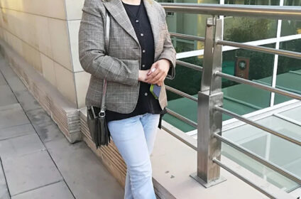 A person standing on an outdoor balcony, wearing a checked blazer, black top, and light jeans, smiling at the camera with buildings reflected in the windows behind.