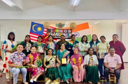Group photo from an international learning and collaboration event featuring educators and participants from different countries, standing and seated on a stage with Malaysian and Indian flags in the background, holding awards and souvenirs in recognition of cross-cultural educational exchange.