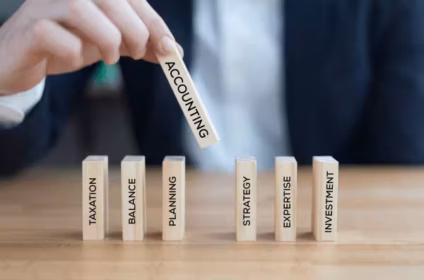 A professional image showing a person in a suit placing a wooden block labeled “ACCOUNTING” among blocks labeled “TAXATION,” “BALANCE,” “PLANNING,” “STRATEGY,” “EXPERTISE,” and “INVESTMENT” on a desk.